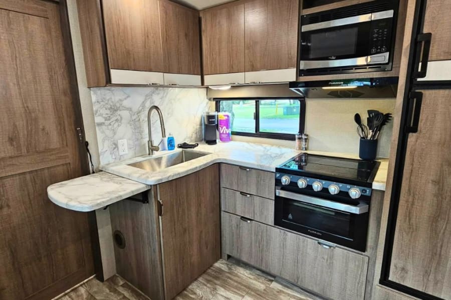 Inside an RV rental, a compact kitchen with wood-toned cabinets, a sink, stove, and microwave, plus an extension counter.