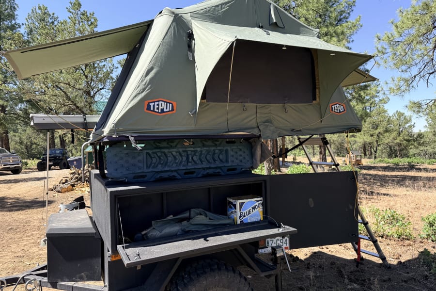 A green Tepui rooftop tent deployed on an overland camping trailer at a sunny campsite. Great for your next rv rental adventure.