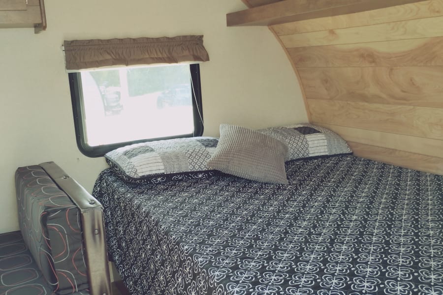 This RV rental's interior shows a cozy bed with patterned linens, a window, and wood-paneled ceiling/wall.