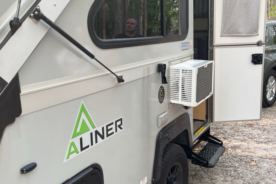 A light-colored A-liner pop-up camper rv rental with an external AC unit, open door, and a person visible inside.