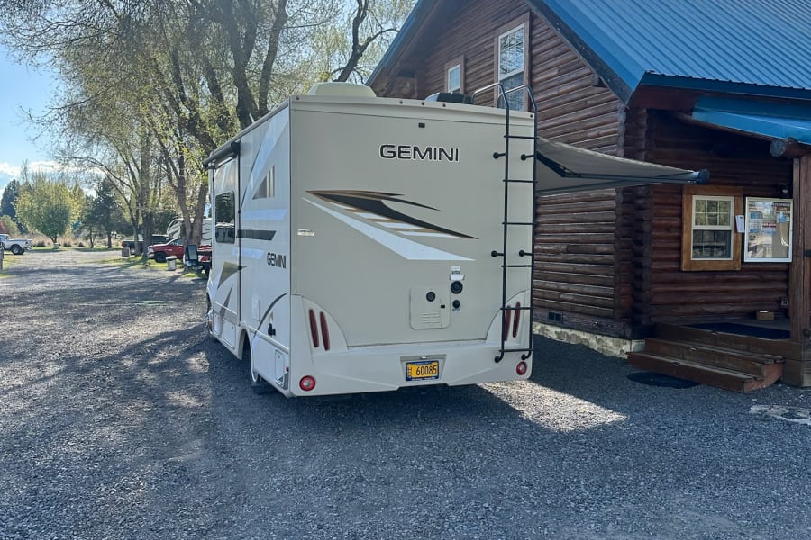 Thor Gemini rv rental on a gravel lot next to a log cabin with a blue roof under bright sunlight.