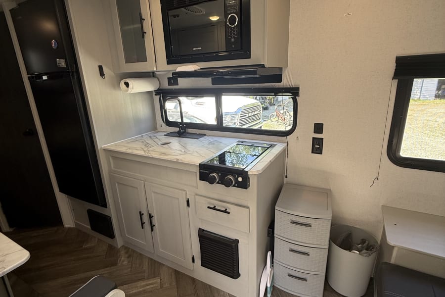 This RV rental features a compact, modern kitchen: microwave, sink, two-burner stove, fridge, cabinets, dinette, and herringbone floor.