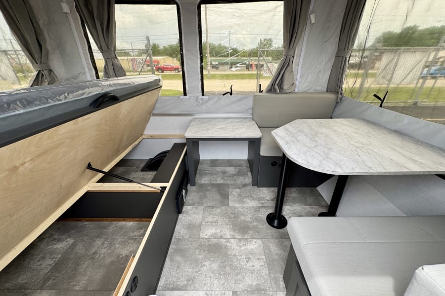 This RV rental features a modern pop-up camper interior with a dinette, tables, cozy seating, and lift-up bed storage.
