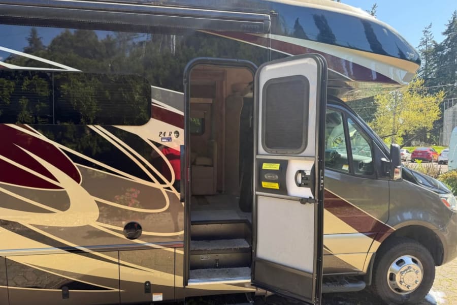 Side view of an RV rental with the passenger door open, revealing interior steps. Features a distinctive dark & gold exterior design.