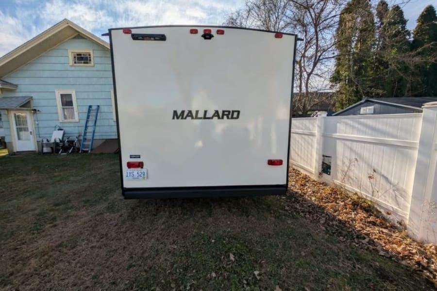 White Mallard RV rental, license plate 1YS 529, parked in a grassy residential yard. A blue house is to the left, a white fence to the right.