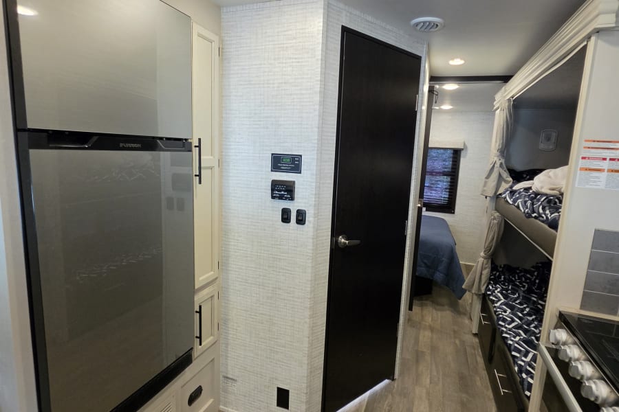 Bright RV rental interior: view of kitchen fridge, control panel, hallway leading to bunk beds & a private bedroom.