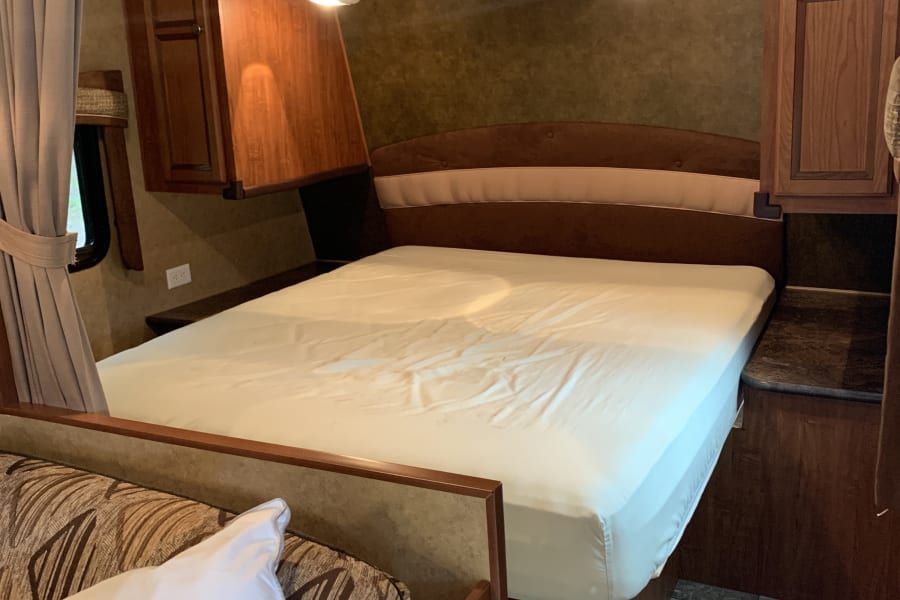 RV rental bedroom: A comfortable bed with white sheets, overhead wooden cabinets, window, and a sofa in the foreground.