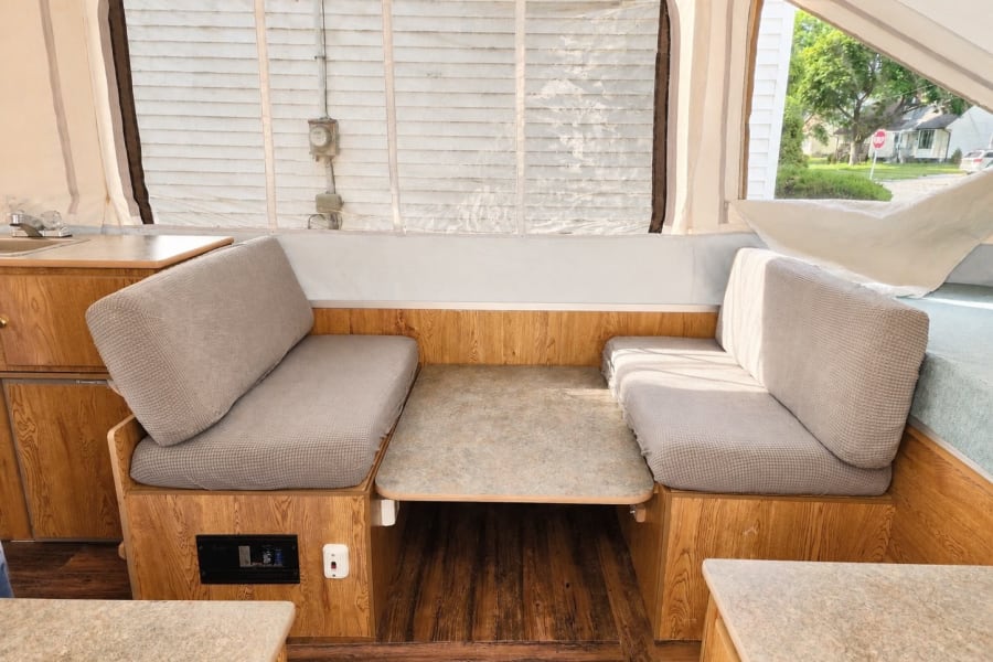 RV rental: Inside a pop-up camper dinette with grey cushioned benches, a table, wooden cabinetry, and a sink area.