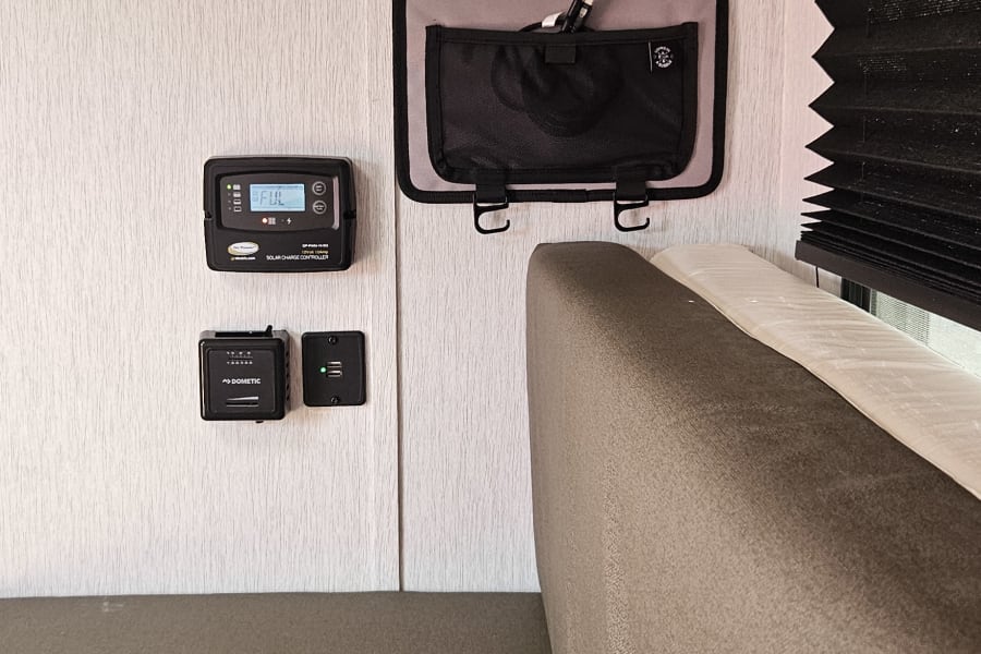 This RV rental interior features a solar controller, Dometic panel, USB port, and fabric storage pouch next to a cushioned bed.