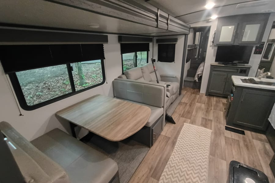 This RV rental offers a modern living space with a dining booth, sofa, compact kitchen, and steps to an upper area.