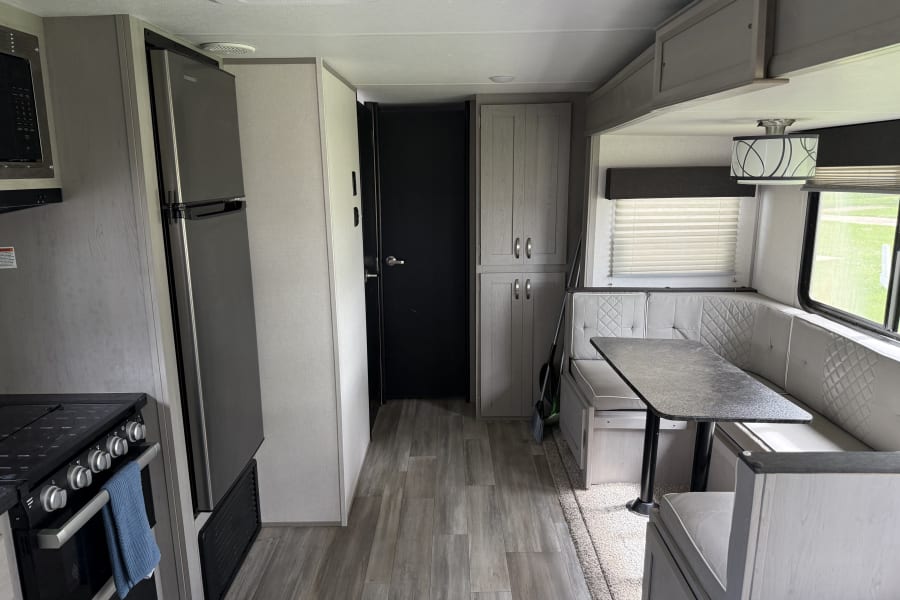 This modern RV rental interior features a kitchen with a fridge and stove, a pantry, and a U-shaped dinette with windows.