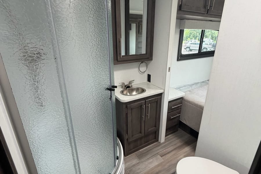 Modern RV rental bathroom: glass shower, sink vanity, toilet, and wood-look floor, with a peek into the bedroom.