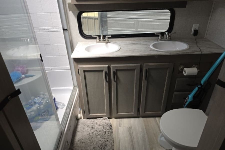 RV rental bathroom showing a double vanity with two sinks, a toilet, and a shower stall with a glass door. Light wood floor and cabinets.