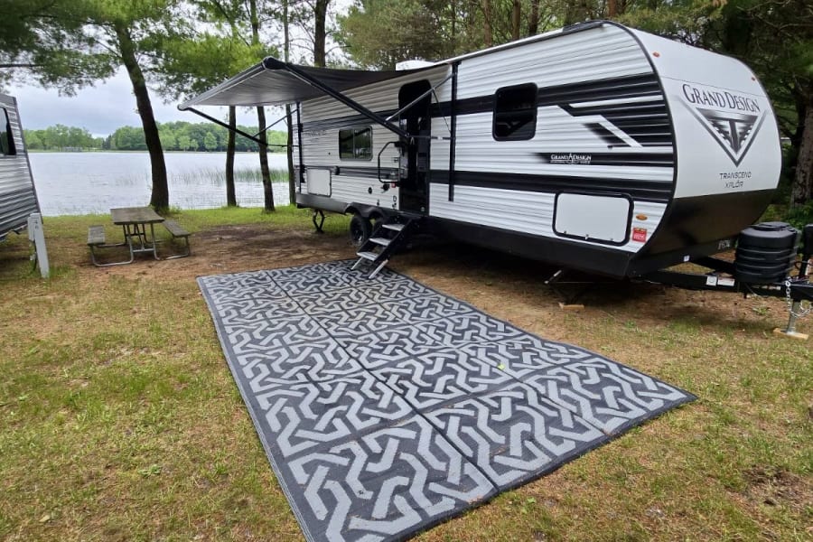 Scenic RV rental campsite: A Grand Design travel trailer with awning & outdoor mat, parked lakeside with a picnic table.