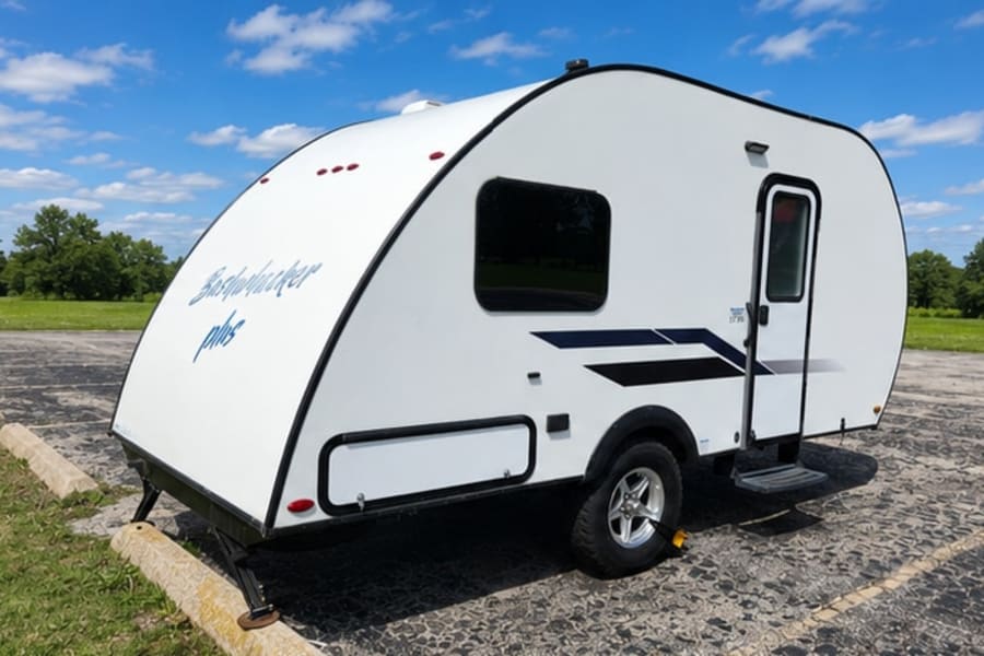 White Bushwhacker plus RV rental: a compact teardrop camper trailer parked on asphalt under a blue sky.