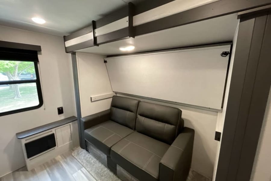 Modern RV rental interior featuring a cozy gray sofa, fold-down bed, window, and small heater unit for compact comfort.
