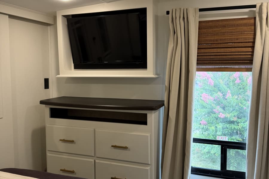 RV rental bedroom with a mounted TV above a dresser, and a window with blinds, curtains, and a vibrant garden view.