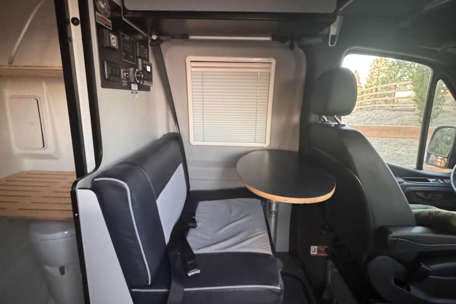 Interior of an RV rental, featuring a dining area with a bench seat and table, driver's seat, and a small toilet.