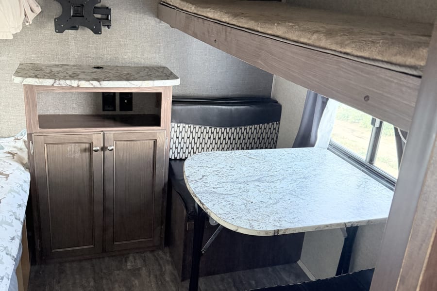This RV rental interior features a functional dinette, an overhead bunk bed, and a cabinet with a wall-mounted TV bracket.