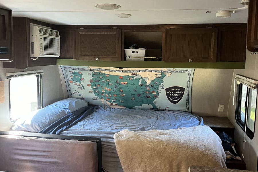 This cozy RV rental interior features a bed, national parks map, AC unit, and a small dining area, perfect for travel.