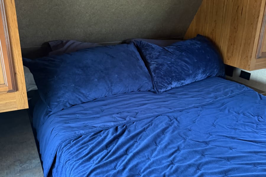 A cozy blue bed with quilted bedding inside an RV rental, featuring warm wooden overhead cabinets and built-in lighting.
