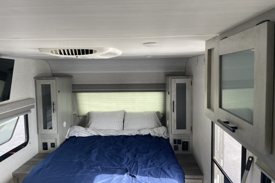 Bright RV rental bedroom with a blue bed, surrounding storage, and an AC unit, providing comfort in a compact space.