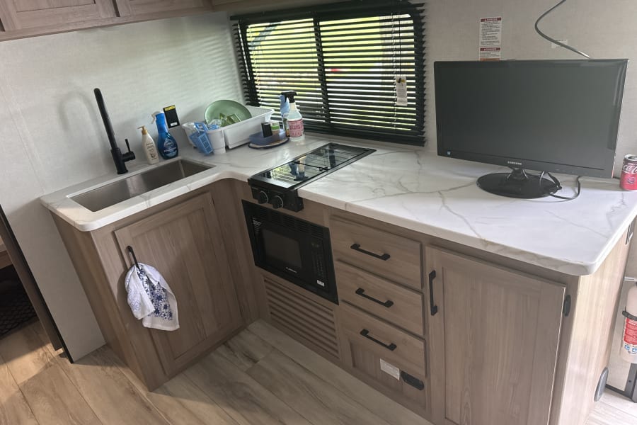 Compact rv rental kitchen with sink, stove, microwave, TV, marble-look counters, and wood cabinets.