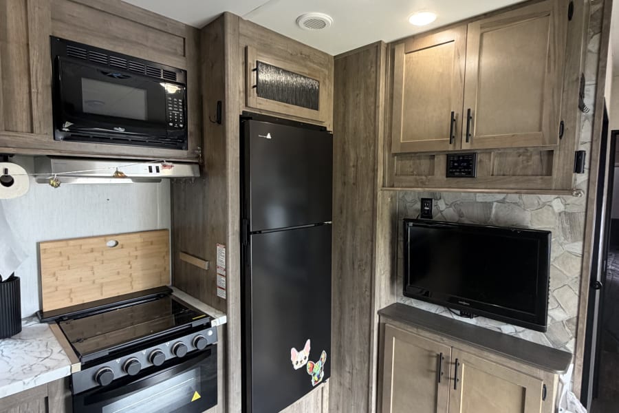 RV rental interior: A modern kitchen area with black microwave, oven/stove, large fridge, and TV, surrounded by wood-grain cabinets and flooring.