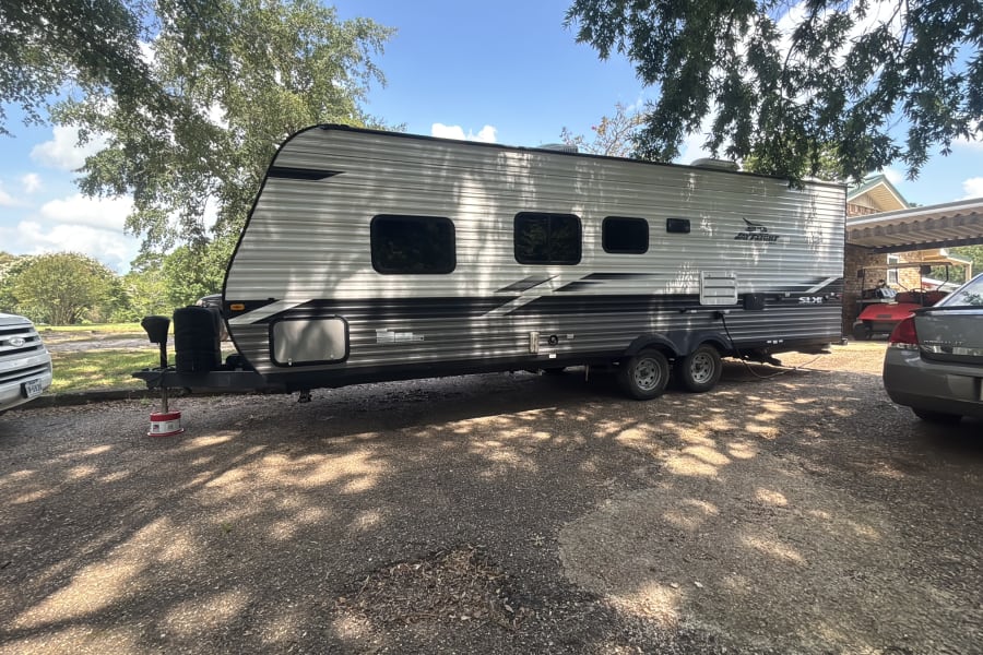 White & grey Jay Flight SLX RV rental parked on a gravel lot under shade trees, with other vehicles nearby.