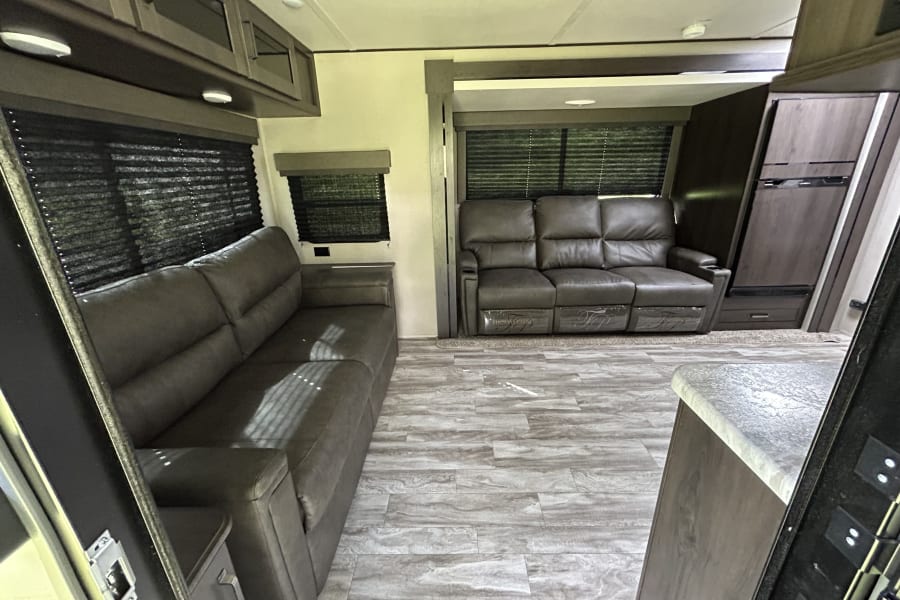 Spacious RV rental living area featuring two comfy sofas, one with recliners, wood-look flooring, and dark cabinetry.