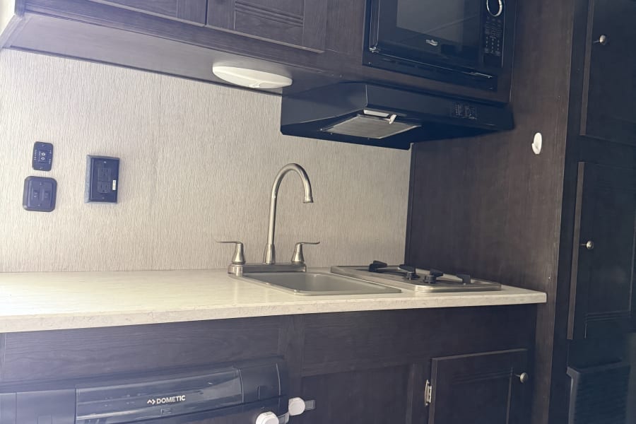 A compact kitchen in an RV rental, featuring dark cabinets, white countertops, a sink, stove, microwave, and Dometic fridge.