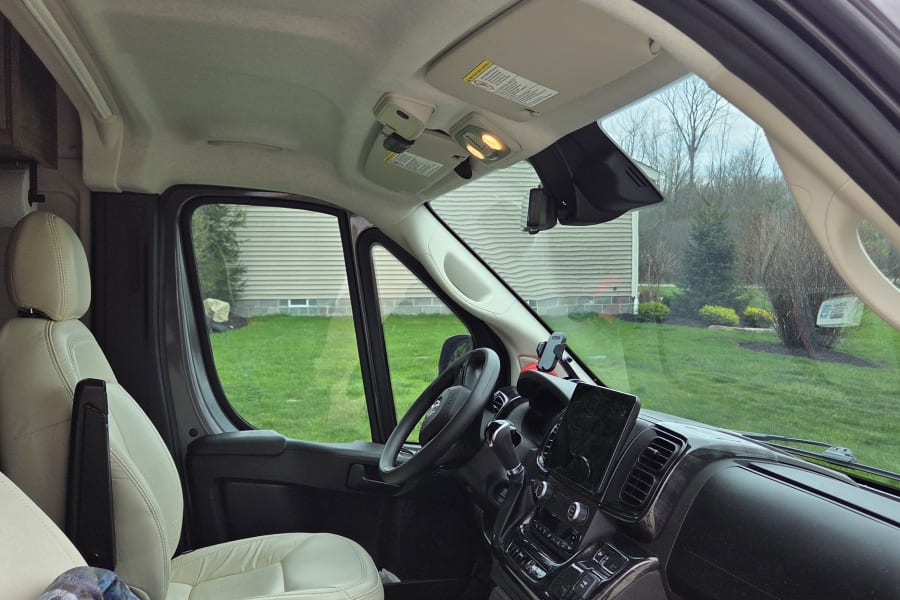 Interior view of an RV rental cockpit featuring light-colored seats, a modern dashboard, and a yard view through the windshield.
