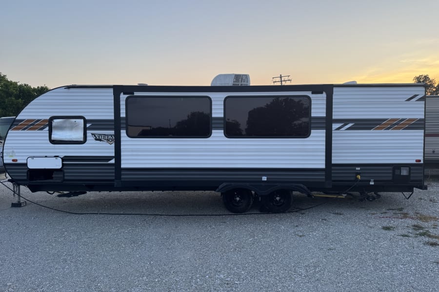 A travel trailer (RV rental) parked on gravel at dusk, featuring windows and an AC unit, ready for your outdoor adventure.