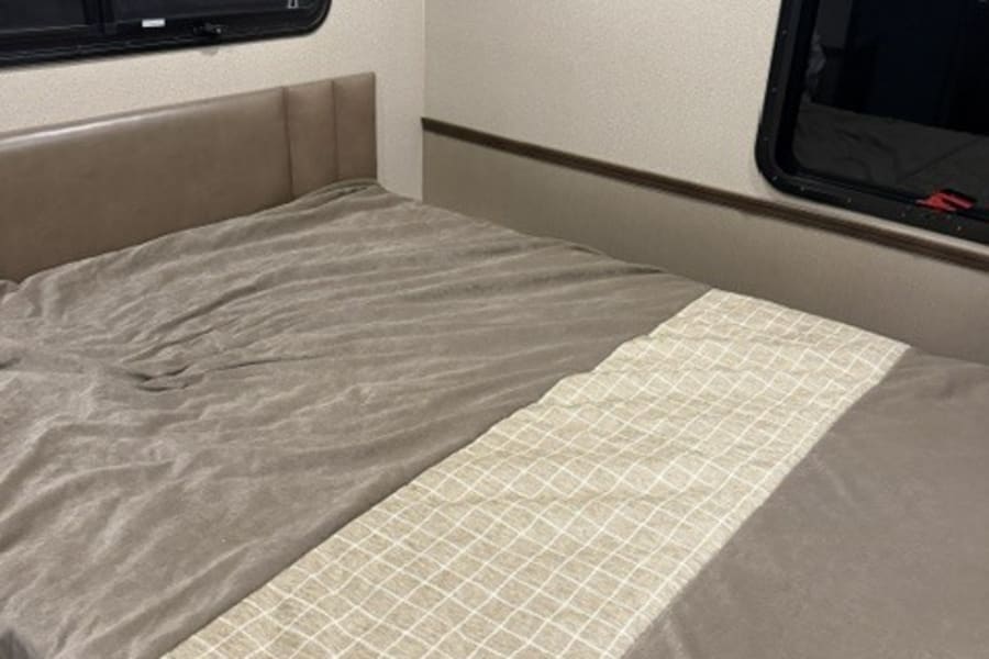 RV rental bedroom: A made bed with neutral bedding, a padded headboard, windows, and overhead cabinets.