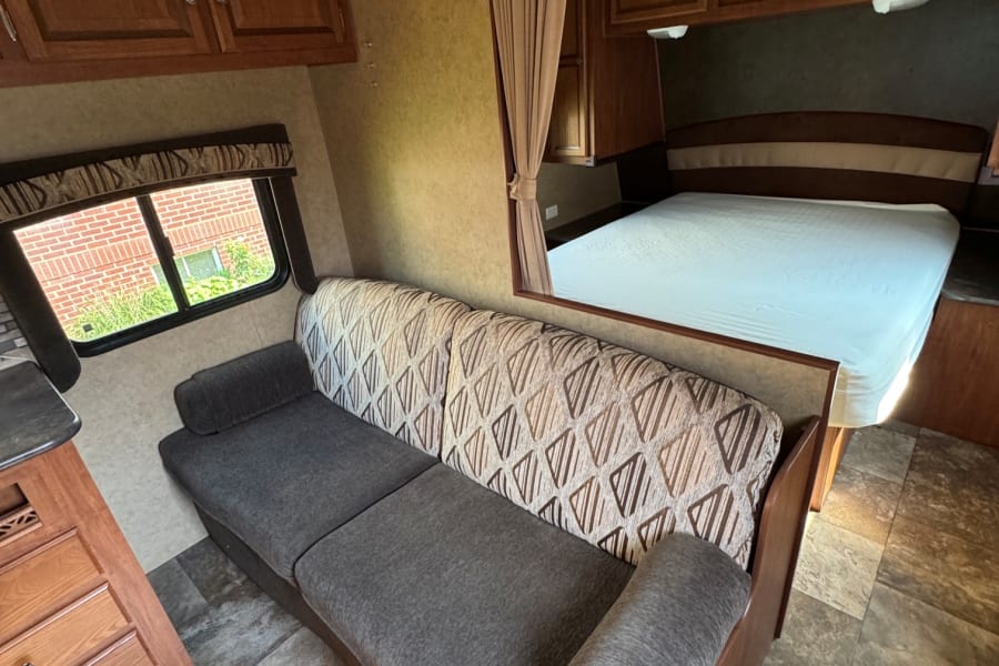 RV rental interior showing a comfortable sofa, a bed with white sheets, and wood cabinetry, with a window view.