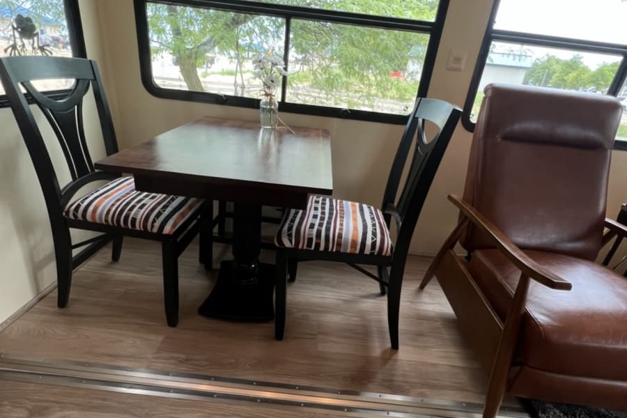 RV rental interior: A cozy dining area with a wooden table, two chairs, and a brown armchair by large windows.