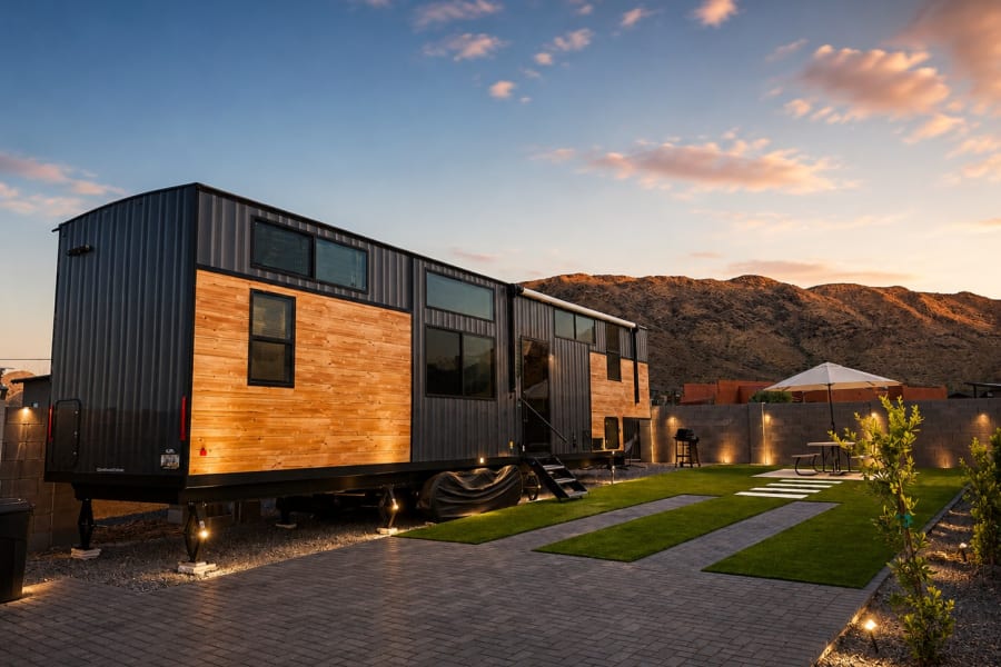 Modern tiny home RV rental with a sleek design, featuring dark metal and wood siding, a landscaped yard, and mountains at sunset.