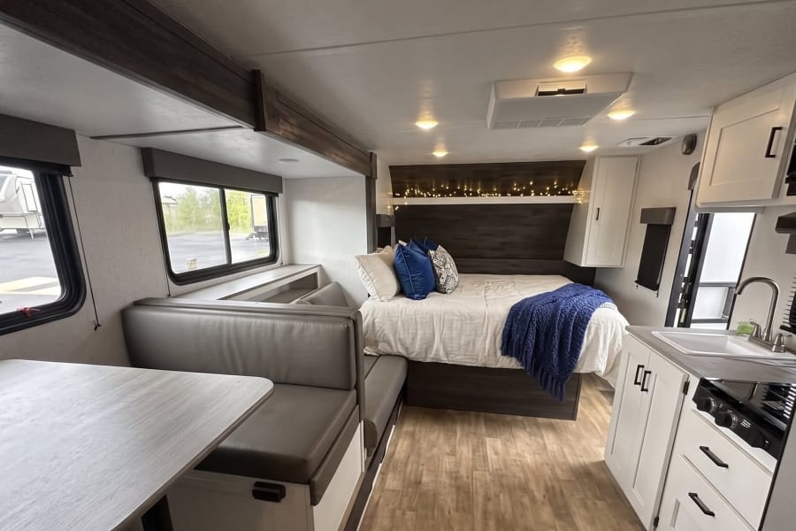 This modern rv rental interior features a cozy bed with string lights, a dining booth, and a compact kitchen area.