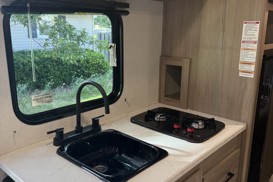 RV rental kitchen interior: modern black sink, stovetop, light counters, wood cabinets & window view.