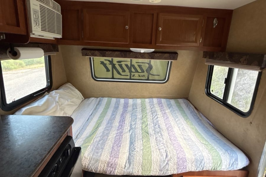 Cozy RV rental interior featuring a comfortable bed, windows, AC, and kitchenette area, ideal for road trips.