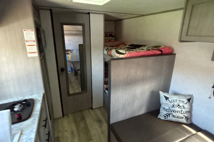 RV rental interior: compact kitchen, mirrored door, cozy bunk bed, and dinette with 'adventure' pillow. Ready for your trip!