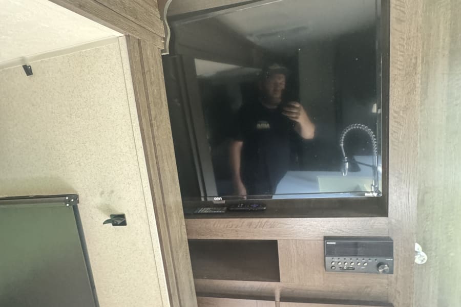 Inside an rv rental: mounted TV, stereo, cabinets. Reflection shows a person and sink, highlighting a functional interior.