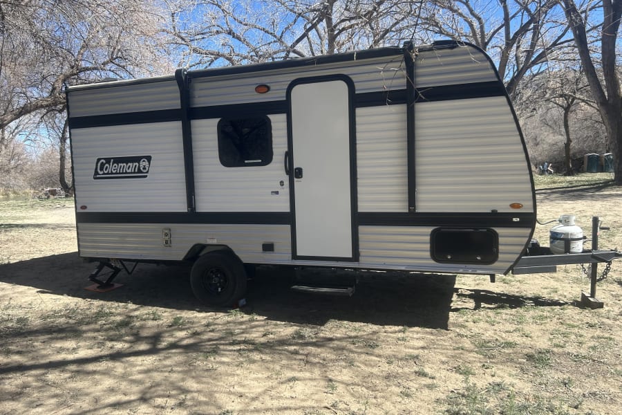 A compact Coleman RV rental, a light-colored travel trailer, is parked on sandy ground under bare trees, ready for camping.