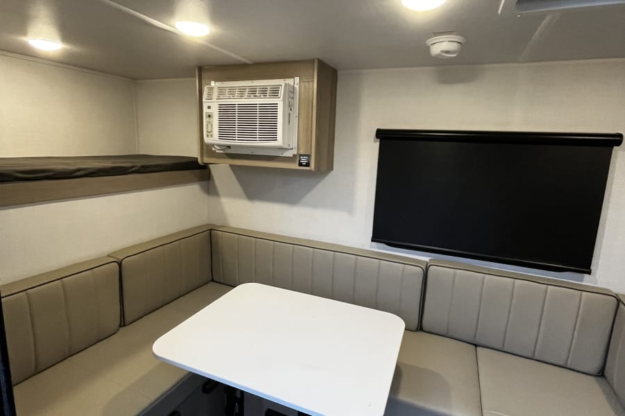 An RV rental interior with a U-shaped dining area, bunk bed, AC, and window blinds.