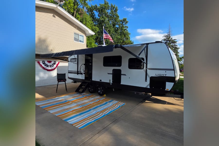 This white Coleman travel trailer, an rv rental, is parked on a driveway with an extended awning, striped mat, and grill setup.