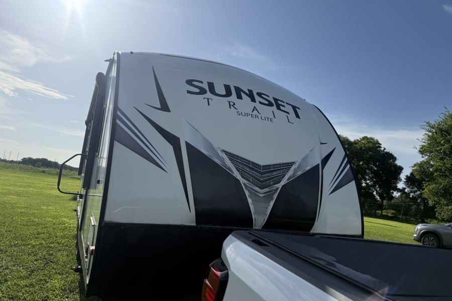Sunny day RV rental: A white and black Sunset Trail travel trailer hitched to a truck, surrounded by green grass.
