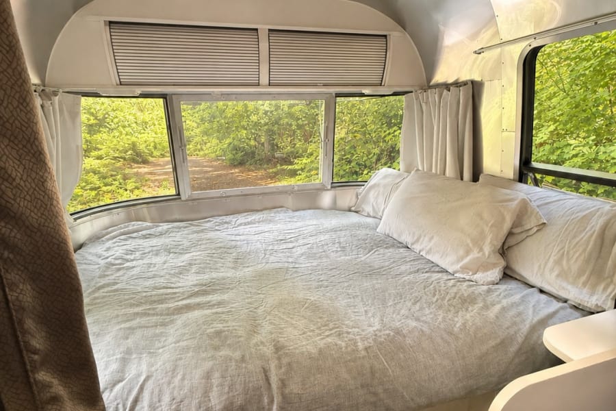 Cozy Airstream rv rental bedroom: comfortable bed, built-in storage, silver interior, and scenic green views from windows.