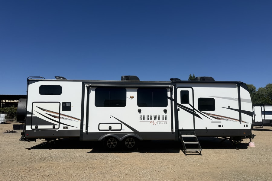 White Rockwood RV rental, a travel trailer with steps, sits on a sunny gravel lot. Ideal for your next outdoor adventure.