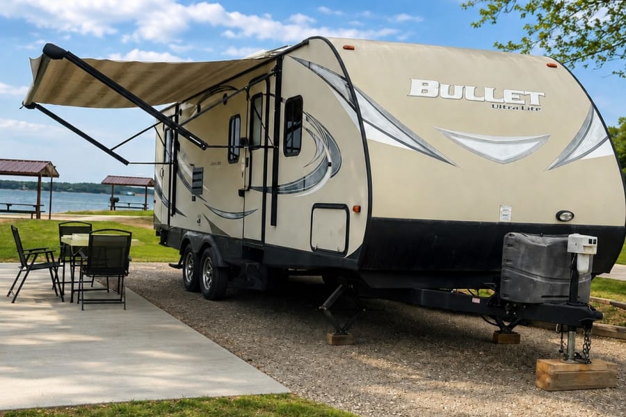 Lakeside RV rental: A tan travel trailer with awning extended at a scenic campsite, ready for your outdoor adventure.