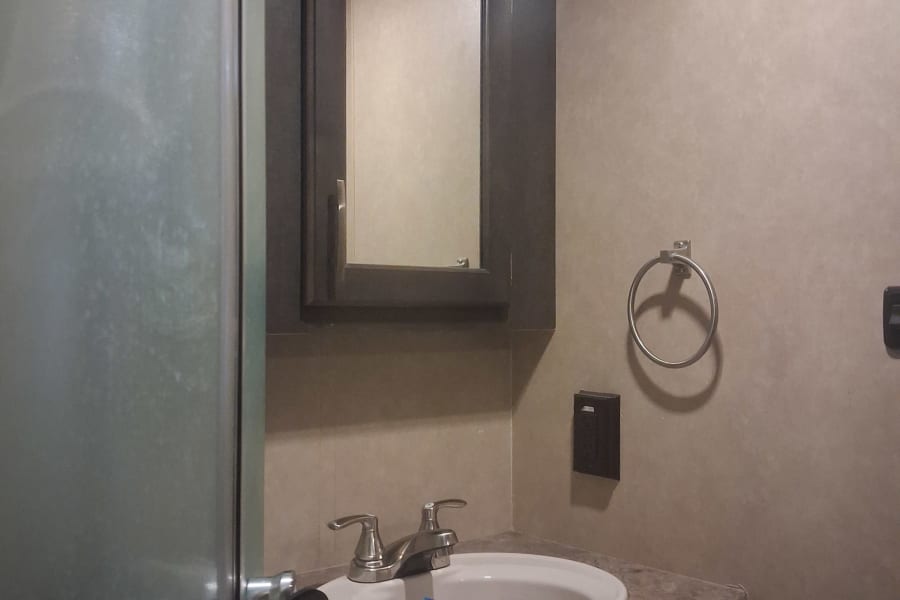 Small, functional RV rental bathroom with a vanity, sink, mirror, and a frosted shower door on the left, offering essential travel comfort.